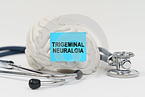 On a white surface next to the stethoscope lies a brain on which a sticker with the inscription - Trigeminal neuralgia