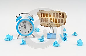 On a white surface there is an alarm clock, crumpled paper and a cardboard sign with the text - Turn Back the Clock