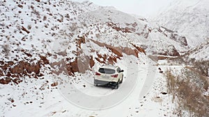 White SUV is driving on a snowy mountain road. Rear and side view. Amazing view of the red mountain canyon Konorchek