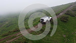 White SUV rides on a mountainous terrain on a dirt road in the cloud. Aerial view.
