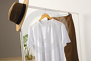 White t shirt, hat and brown trousers hanging from clothes rail with copy space on white background
