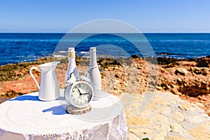 White table with antique clock placed on the rocks of the beach, with sea background