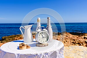 White table with antique clock placed on the rocks of the beach, with sea background and intense blue sky