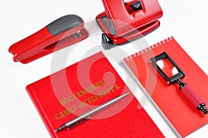 On a white table closeup are red office accessories: stapler, hole punch, Notepad, magnifier, book and pen