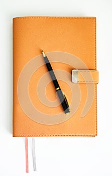 An orange notebook with a magnetic clasp sits on a white table with a black pen on it.