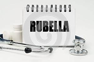 On a white table lies a stethoscope, pills and a notebook with the inscription - RUBELLA