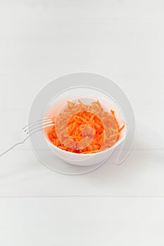 On the white table is a plate with grated fresh carrots and a fork. copy space.