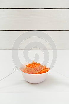 On the white table is a plate with grated fresh carrots and a fork and knife. Wooden background