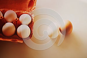 On a white table there is an open orange cardboard package with white chicken eggs, and next to it are two eggs. Healthy nutrition