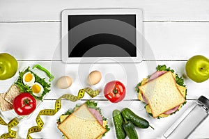 White tablet computer with a blank screen on the kitchen table with sandwiches, fresh vegetables, bottle of water and