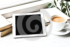 White tablet sits on a table next to a cup of coffee and a stack of books.