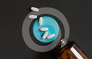 White tablets pills spilling out of a toppled brown bottle over dark background