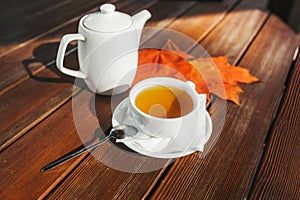 White tea cup with black tea, white teapot on wooden table and autumn maple leaves