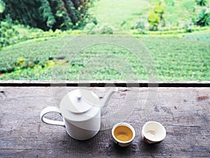 White teapot and cups on vintage wooden table over green tea farm background