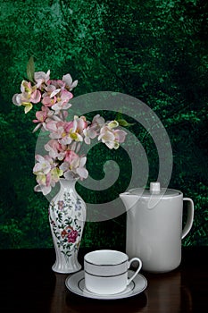 White Teapot and Teacup and Saucer with Artificial Flowers in a White Decorated Porcelain vase on a Mottled Background and Wooden