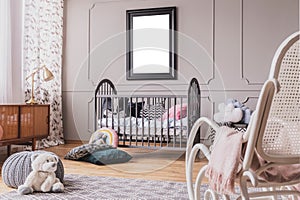 White teddy bear on the floor of stylish baby room interior with grey wooden crib with pillows, white rocking chair and mockup