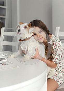 Teenage girl 10 years old sits at the kitchen table and hugs jack Russell`s white furred dog photo