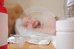 White thermometer on a pack of pills and two bottles with medicine in focus, sick man sleeping in bad out of focus in the