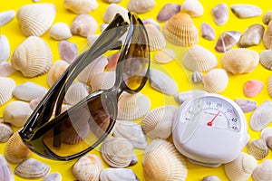 White thermometer with a temperature of +26 degrees Celsius, sunglasses and a lot of different seashells on a yellow background