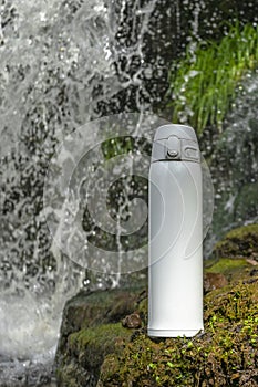 White thermos of tea or coffee on stone near waterfall with splashes of water drops. Adventure hiking tourism concept