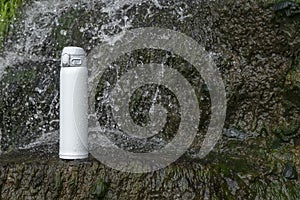 White thermos of tea or coffee on stone near waterfall with splashes of water drops. Adventure hiking tourism concept