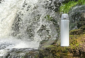 White thermos of tea or coffee on stone near waterfall with splashes of water drops. Adventure hiking tourism concept