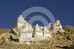 A white Tibetan Buddhist stupas temples on a deserted stone mountainside against the backdrop of a clean dark blue sky and moon