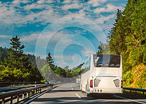 White tour bus or coach on a two lane motorway travelling through a tree lined hilly countryside on a sunny day