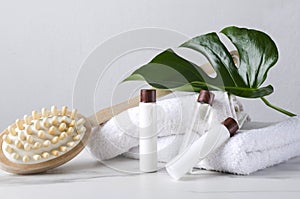 White towels, wooden brush for massage, bottles of lotion, body cream, green tropical leaf as a decor on the white table against b