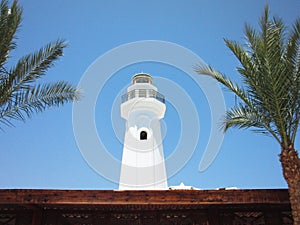 The white tower of minaret on a blue sky background between two palms