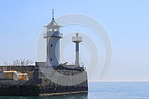 The white tower of a small old lighthouse on a rocky promontory. In the background is a calm blue sea and sky. Yalta