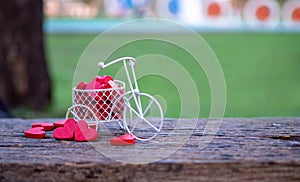 White toy bike carrying red wood hearts. Red wood hearts fall on the wooden floor, green background of grass. Heart-shaped toys