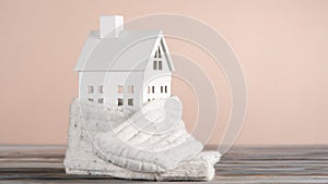 White toy model of house with white knitted scarf made of carton paper on grey table on pink background. Heating system.