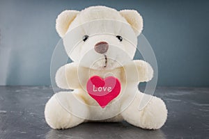 White toy teddy bear with heart on a gray background. The symbol