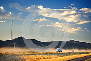 A white truck on a dusty freeway. In the background are dark brown hills and a dark blue sky with fluffy clouds. A mobile phone to