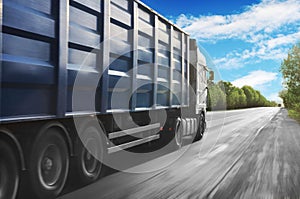 White truck in motion with a blue container driving on the countryside road against blue sky with white clouds