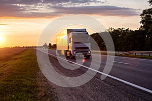 A white truck with a semitrailer transports cargo on the highway in the evening against the backdrop of sunset. Compliance with
