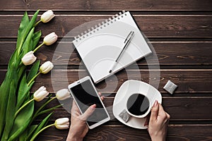 White tulips on a wooden table with an empty notebook, smartphone and a Cup of coffee in women`s hands