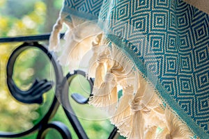 A white and turquoise Turkish peshtemal / towel on a wrought iron railings with blurry nature in the background.