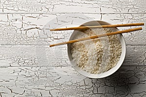 White uncooked, raw long grain rice in white bowl with chopsticks on rustic white wooden table with copy space top view flat lay