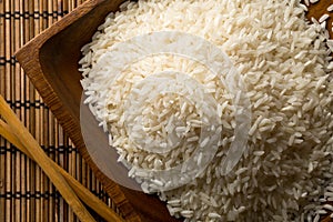 White uncooked, raw long grain rice in wooden bowl with chopsticks on bamboo mat top view flat lay from above