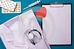 White uniform of a doctor and a stethoscope lie on the table, next to a red heart, pills