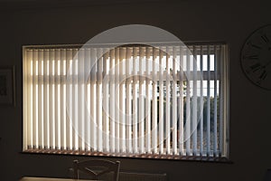 White vertical blinds on a window with bright sunlight behind them. there are slightly open