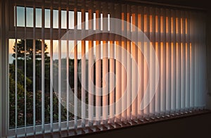 White vertical slat blinds hanging in front of a window as the sun is setting turning the light golden. photo