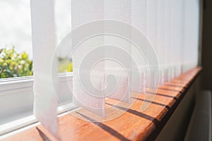 White vertical window blinds slats with cordless glued weighted pockets on the end casting shadows on the wooden window sill