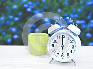 white vintage alarm clock and green cup of coffee on the table in the garden with purple flowers background