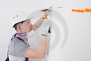 In the white wall, holes are made and orange dowels are hammered with a hammer, a worker in a protective helmet