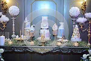 White wedding cake decorated by flowers standing on festive table with lots of snacks on side. Violet flowers on foreground.