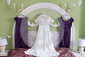 White wedding dress and two lilac bridesmaidâ€™s dresses on a hanger on the wall. Morning of the bride. Fees.