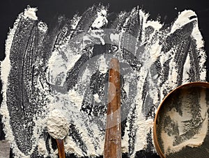 White wheat flour scattered on a black table and very old brown wooden rolling pin, round sieve